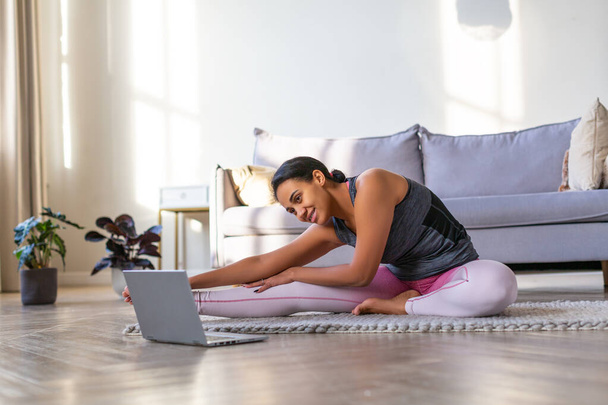 自宅でオンラインで動作します。アフリカ系アメリカ人女性がノートパソコンのモニターの前で家で運動している。伸び. - 写真・画像