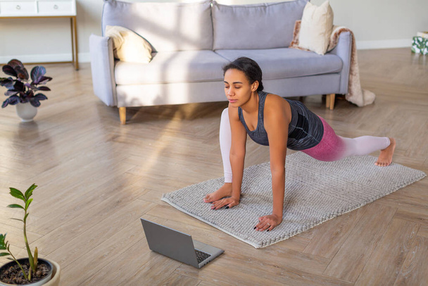 Estiramiento en casa en línea. Mujer afroamericana ejercitándose en casa frente a un monitor portátil. - Foto, imagen