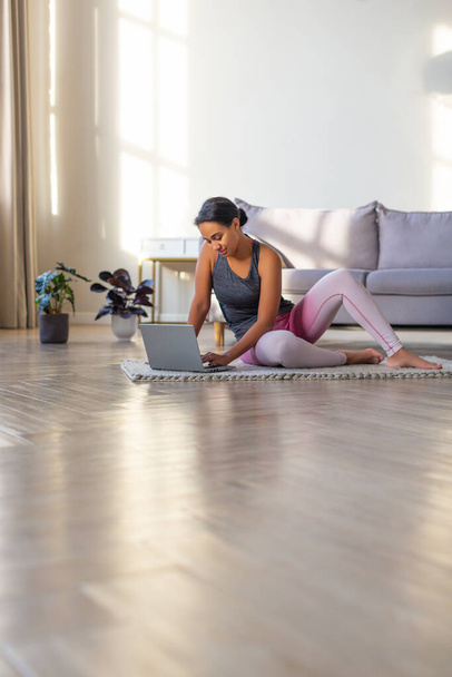 Smiling African-american woman preparing for a workout at home in front of a laptop monitor. Vertical photo. - Foto, afbeelding