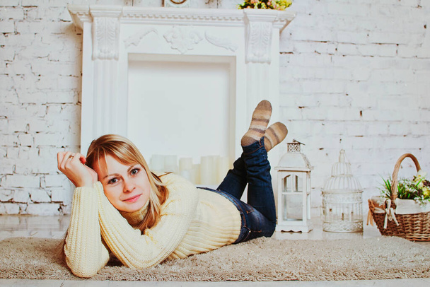 Junge blonde attraktive Frau in Jacke und Jeans sitzt im Winter zu Hause am weißen Kamin. - Foto, Bild