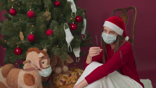 Een meisje met een medisch masker heeft sterretjes bij een kerstboom. Vakantie pandemie. - Video