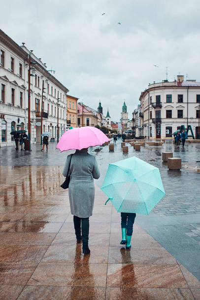 Back view of mother and her daughter holding the pink and blue umbrellas walking in a downtown on rainy gloomy autumn day - Φωτογραφία, εικόνα