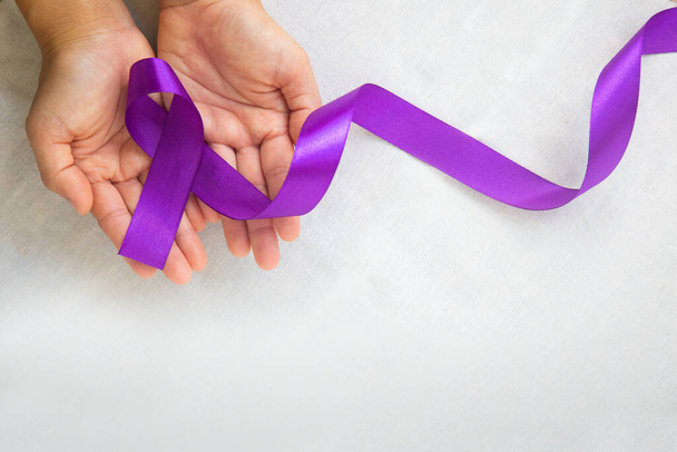 Manos sosteniendo cinta púrpura o violeta en tela blanca con espacio para copiar. Cáncer de páncreas, conciencia sobre el cáncer testicular, sobreviviente de cáncer, leiomiosarcoma, Día Mundial del Cáncer. Asistencia sanitaria, concepto de seguro. - Foto, Imagen