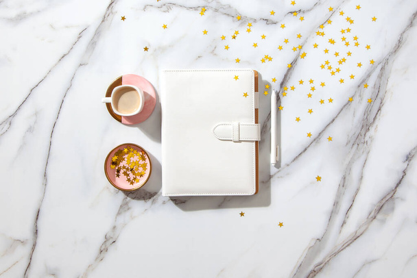 Notebook with empty pages for your text, pen, coffee cup, milk, small gift boxes with pink ribbon and golden sparkles on white marble table. New Year and Christmas concept, top view. Festive holidays - Fotó, kép