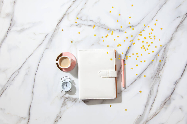 Notebook with empty pages for your text, pen, coffee cup, milk, small gift boxes with pink ribbon and golden sparkles on white marble table. New Year and Christmas concept, top view. Festive holidays - Фото, изображение