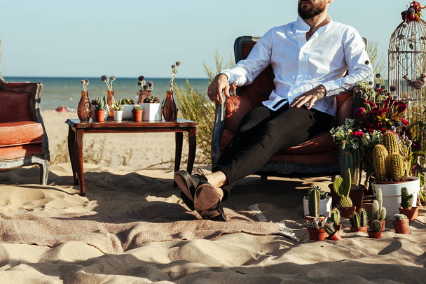 Обрезанное фото жениха, сидящего на стуле на свадебной церемонии на пляже. расслабленный мужчина в уютной обстановке для свидания. свадебное убранство с кактусом и суккулентами на берегу моря. - Фото, изображение