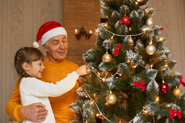 Grandfather and granddaughter decorating xmas tree, looking at fir tree with smile, child in white jumper, senior man wears yellow shirt and santa claus hat, posing indoor in festive room. - Foto, Imagem