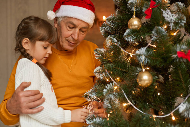 Family celebrating Christmas at home, grandad and grandchild decorating christmas tree together, look concentrated, wearing casual clothing, mature man in santa claus hat. - Фото, изображение
