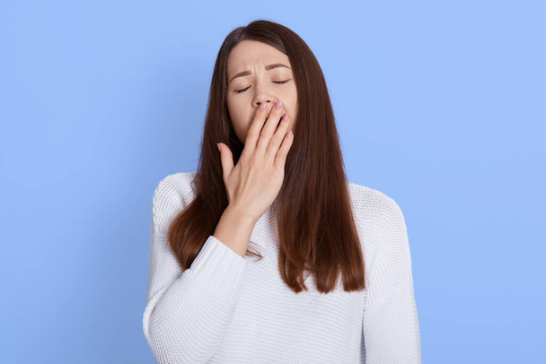 Young beautiful woman yawning, showing tired gesture, covering mouth with her hand, keeping eyes closed, having long dark hair, wearing white casual shirt, posing isolated over blue background. - Foto, Bild