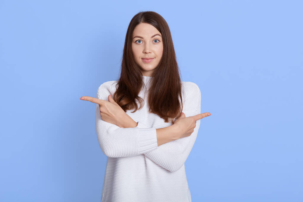 Young beautiful woman wearing casual white shirt over blue background, pointing to both sides with fingers, different direction disagree, looks at camera, choosing where to go. - Фото, зображення
