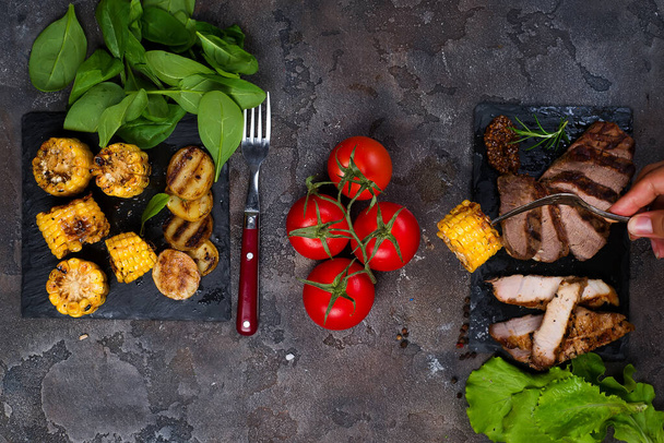 female hand holds a fork with grilled corn on the background fresh three types of grilled steak on slate plate with spinach, tomato and grilled potatoes on stone background with copy space, flat lay - 写真・画像