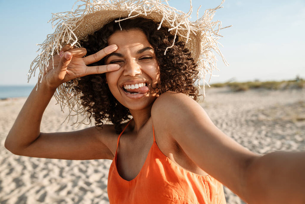 Image of positive young african woman taking a selfie by camera and showing peace gesture at the beach outside - Fotó, kép
