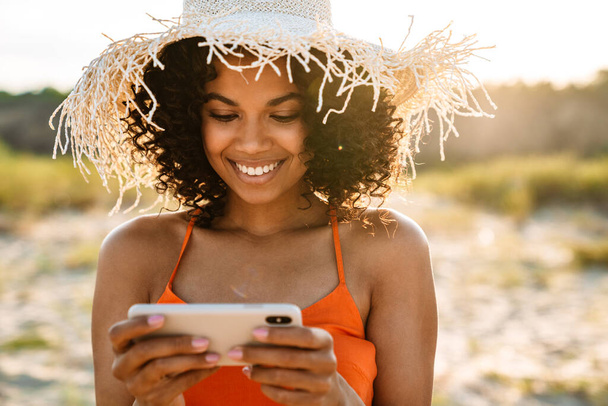 Image of pretty cheery young african woman watching videos by mobile phone at the beach outside - Фото, зображення