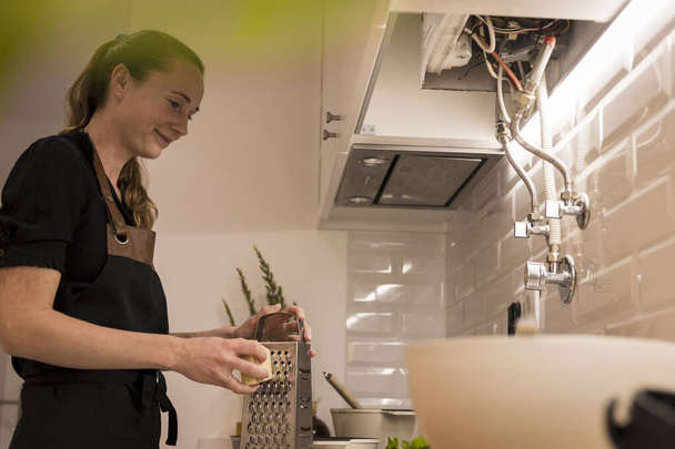 Młoda piękna kobieta stoi przy piecu i tarcie sera dla zdrowego przepisu żywności. Urocza Norweżka pracująca jako bloger kulinarny i trener stylu życia przygotowująca jedzenie w kuchni - Zdjęcie, obraz