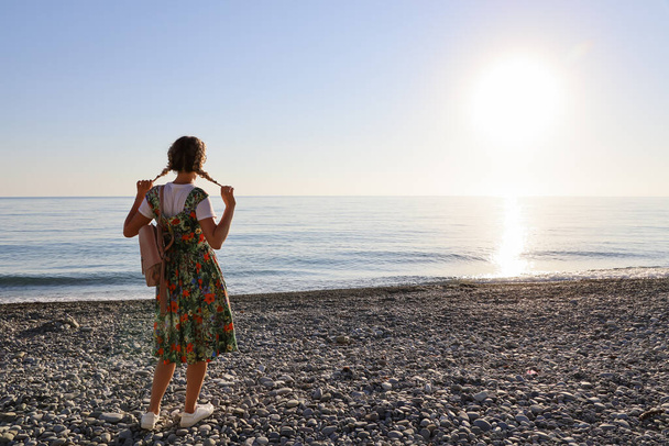 Jeune femme avec des nattes se tient avec son dos seul sur le bord de mer pierreux au coucher du soleil avec espace de copie - Photo, image