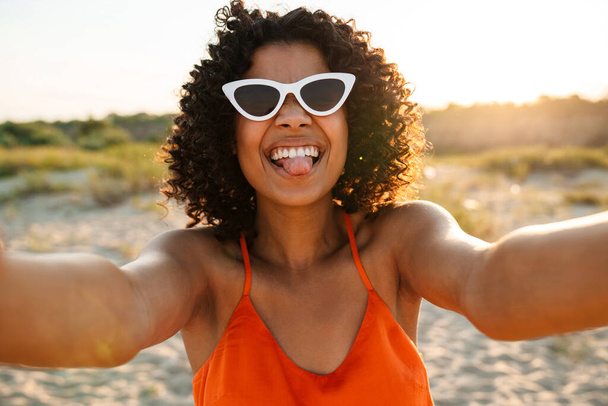 Image of pretty smiling young african woman taking a selfie by camera at the beach outside - Фото, изображение