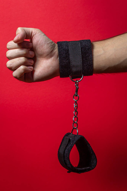 Soft handcuffs. Male hand in soft handcuffs on a red background. BDSM accessories - Zdjęcie, obraz