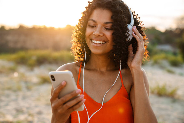 Happy young african woman listening to music with headphones at the beach, holding mobile phone - Foto, immagini