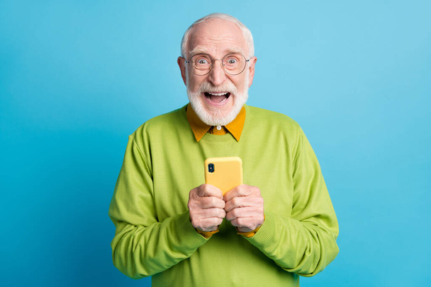 Photo of retired old man hold telephone open mouth crazy face wear specs green pullover isolated blue color background - Fotoğraf, Görsel