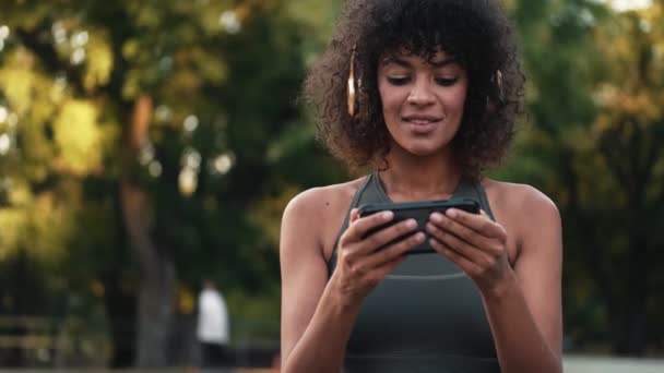 Young african fitness woman using mobile phone outdoors in park while listening music in headphones - Filmmaterial, Video