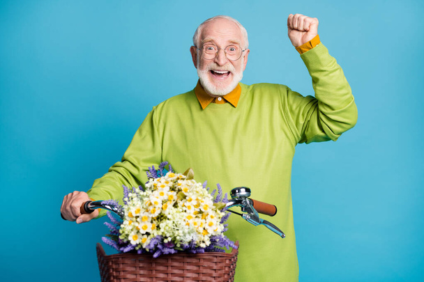 Photo of pensioner old man hold bike flower basket raise fist open mouth wear glasses green sweater isolated blue color background - Фото, зображення