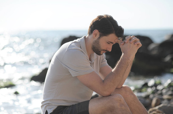 Depressed sad young man sitting on a sea beach, palms on face - Фото, изображение