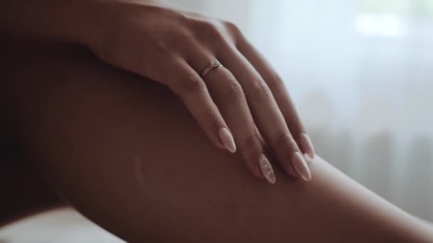 Kavkazská dívka s úhlednou manikúrou a prstenem jemně hladí prsty nohou elegantně ohnutou v koleni, sedící na posteli proti oknu a se sklem v druhé ruce a krátkým bílým kabátem - Záběry, video