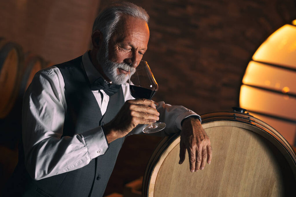 Senior man who could be a winemaker, a sommelier, a connoisseur or simply a wine lover, holds up a glass of red wine and sniffs the bouquet appreciatively. Black background with copy space. - Photo, Image