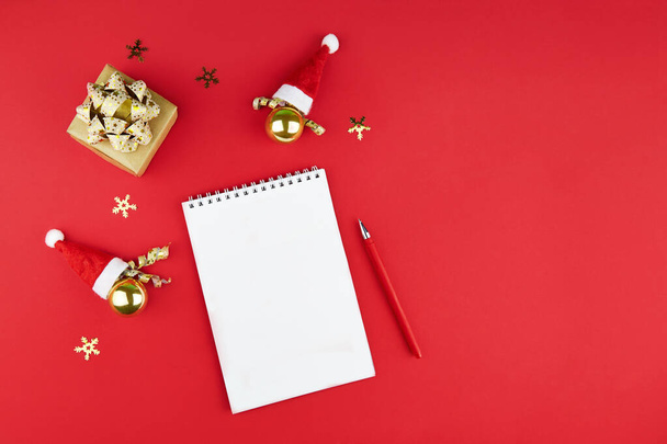 Christmas composition, wish list concept. Notebook with pen, gift, decoration and hats Santa Claus on red background. Flat lay, top view, copy space - Foto, imagen