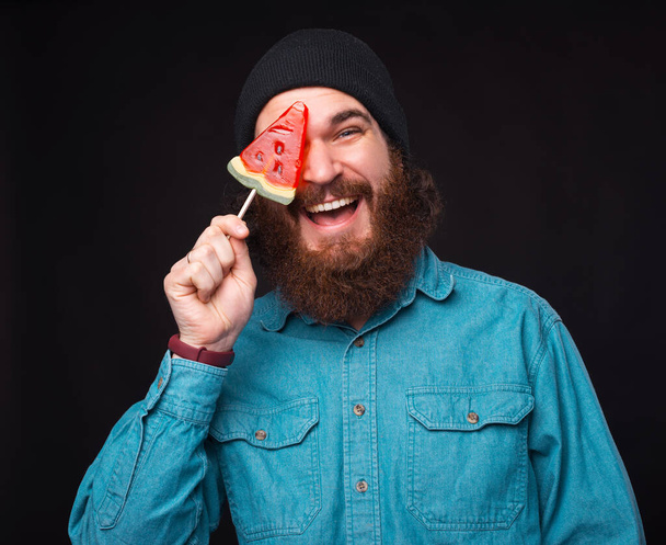 Щасливий бородатий хіпстер покриває одне око паличкою з цукерок кавуна
 - Фото, зображення