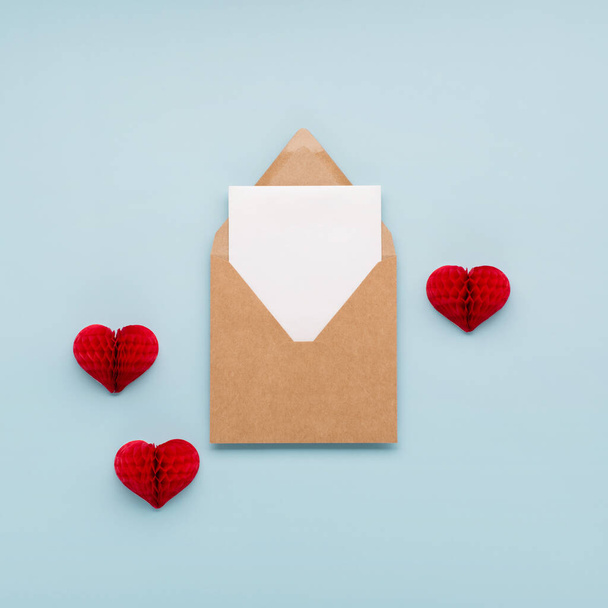 バレンタインデーの休日カード。パステルブルーの背景に封筒付きの赤い小さな紙の心と空のカード。バレンタインデーのコンセプト。フラットレイアウト、トップビュー、コピースペース.  - 写真・画像