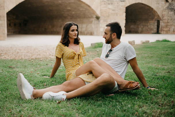 Μια μελαχρινή με κίτρινο φόρεμα κάθεται στα πόδια του φίλου της στο γρασίδι στην παλιά ισπανική πόλη. Μερικοί τουρίστες σε ραντεβού στο πάρκο της Βαλένθια.. - Φωτογραφία, εικόνα