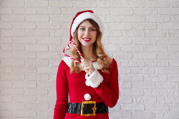 Beautiful smiling young woman wearing sexy santa claus suit for Christmas. Attractive brunette female in tight red dress, new years eve. Copy space background, close up, decorative white brick wall. - Фото, зображення