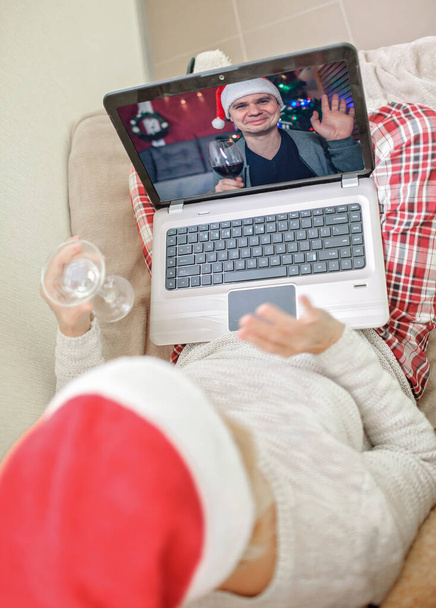Turvallinen online-juhla. Vanhempi nainen, jolla on lasillinen viiniä ja juhlii joulua perheensä kanssa käytännössä internetin ja muistikirjan kautta kotona. Videopuhelu. Kaukainen loma, sisätilojen elämäntapa - Valokuva, kuva