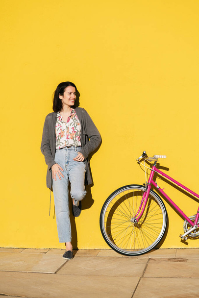 vertical photo of a happy young girl leaning against a colorful yellow wall next to her pink retro bike, concept of active lifestyle and sustainable mobility, copy space for text - Photo, image
