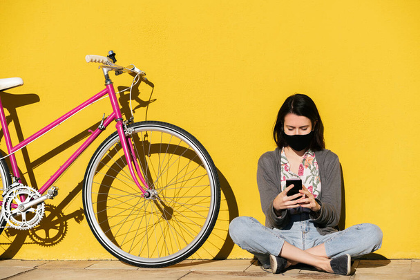 young girl wearing protective mask looking her smart phone sitting next to her bike leaning against a yellow wall, concept of technology, protection against covid and sustainable mobility - Zdjęcie, obraz