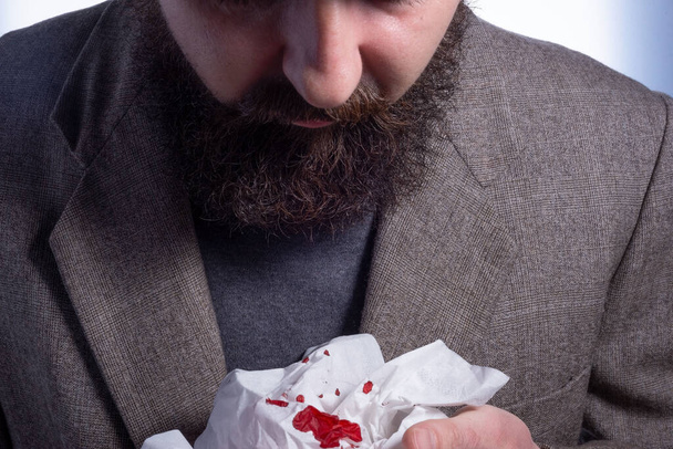 tos sangrienta, hombre de negocios barbudo tosiendo sangre en una servilleta, primer plano, concepto médico cerca de un hombre con tejido ensangrentado - Foto, Imagen