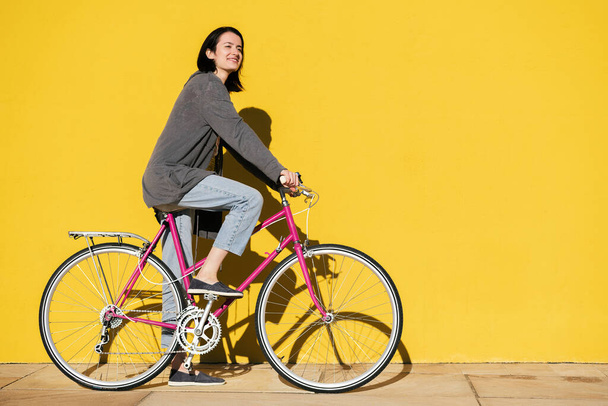 happy young girl smiles riding her retro pink bike next to a colorful yellow wall, concept of active lifestyle and sustainable mobility, copy space for text - Фото, изображение