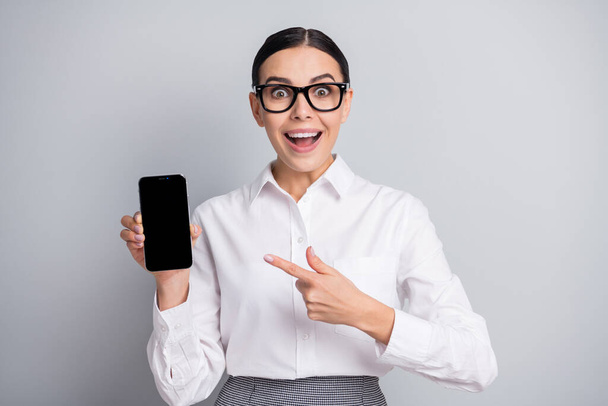 Photo of excited girl hold phone indicate finger screen empty space open mouth wear specs white shirt isolated grey color background - Zdjęcie, obraz