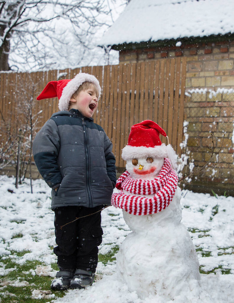 感情的に叫んで幼児の男の子と雪だるまサンタの帽子と鮮やかな赤いストライプのスカーフを身に着けている。冬の休日の気分季節の野外活動気楽な子供時代 - 写真・画像