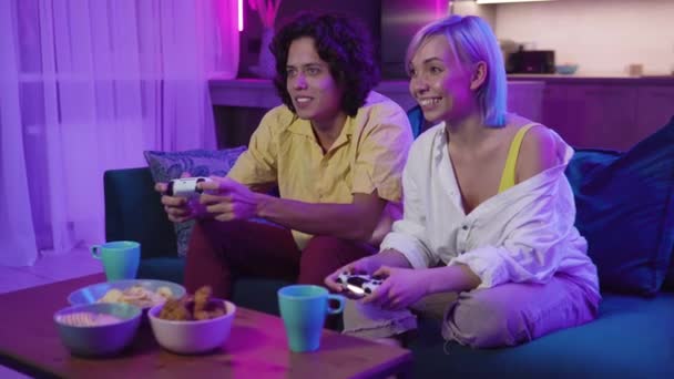 Onnellinen poikaystävä ja tyttöystävä pelaa videopelejä ja pitää joysticks kotona. Millennial pari nauttii pelistä istuessaan sohvalla ja viettää vapaa-aikaa. Suhteen käsite - Materiaali, video