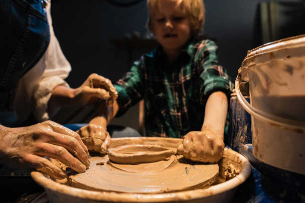 een kleine jongen beeldhouwt een bord uit klei onder toezicht van een leraar in een pottenbakkerij. - Foto, afbeelding