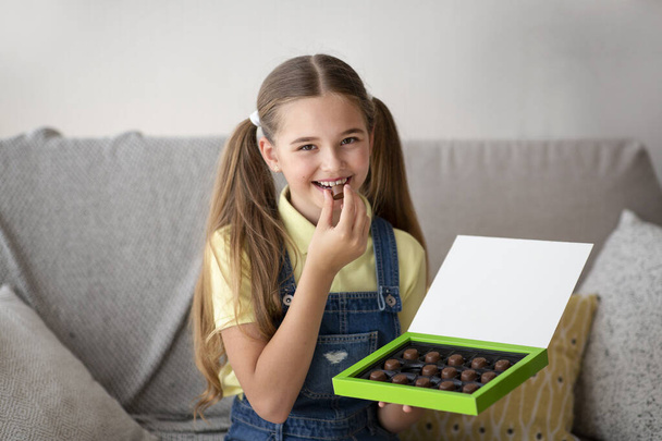 Çikolata yiyen bir kız evdeki koltukta oturuyor. - Fotoğraf, Görsel
