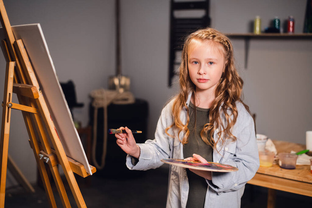 Ένα μικρό κορίτσι ζωγράφος ποζάρει και χαμογελά στην κάμερα κοντά στη ζωγραφική της στο στούντιο καλλιτεχνών. - Φωτογραφία, εικόνα