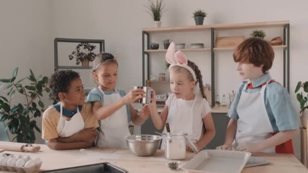 Multiethnic male and female school friends wearing aprons talking, smiling, cooking, standing at table in kitchen. Girl with bunny-ears headband pouring flour through sifter, kids playing with it - Footage, Video