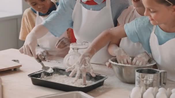 Обрезанные разнообразные дети в белых фартуках, играющие с мукой, наливая ее на стол, от металлической миски до хлебопекарного листа - Кадры, видео