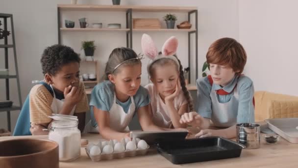 Brust auf von vier verschiedenen Schulkindern in Schürzen, die sich über den Küchentisch beugen, auf Tablets schauen, online nach Rezept suchen. Mixed-Race-Mädchen greift nach Mehlsiebmaschine, rothaariger Junge nimmt es zurück - Filmmaterial, Video