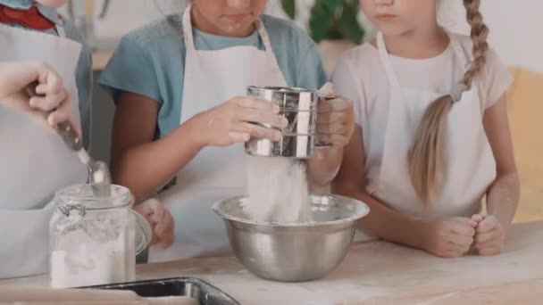 Nahaufnahme eines nicht wiederzuerkennenden Kindes, das mit einem Löffel Mehl aus einem Glas schöpft und es durch einen Sieb in den Händen eines kleinen Mädchens zu einer metallischen Schüssel hinzufügt. Kinder in Schürzen stehen am Küchentisch und kochen  - Filmmaterial, Video