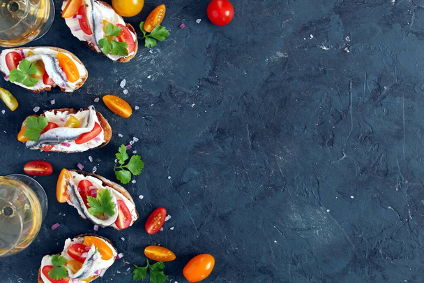 Bruschetta with light cheese, tomato and anchovy served with white wine on dark background. Top view with copy space. Healthy snack. - Photo, image