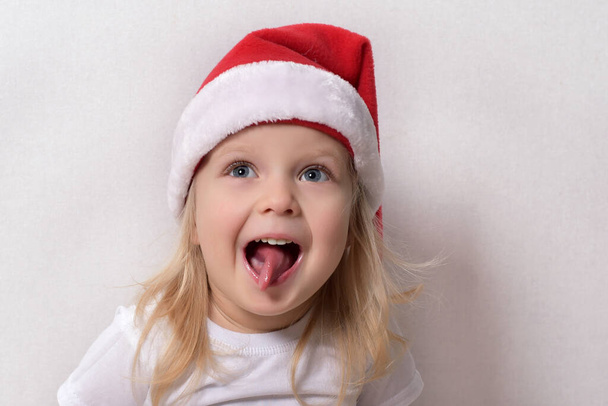 A small child in a Santa hat on a white background with his tongue hanging out - Zdjęcie, obraz